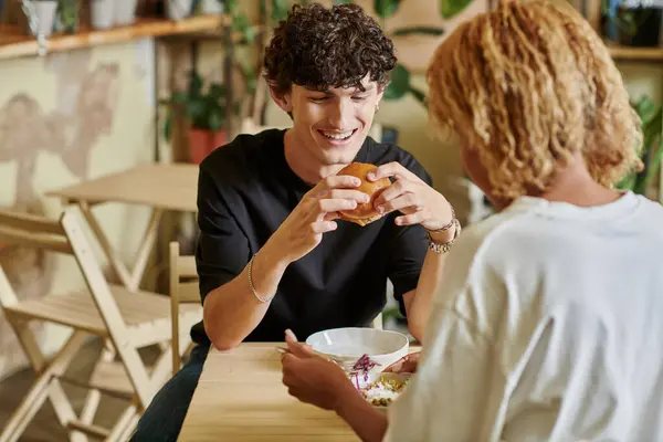 Довольный и кудрявый мужчина ест бургер с тофу рядом с африканской американской девушкой, наслаждаясь салатом в веганском кафе — стоковое фото