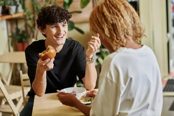 Sorrindo e encaracolado homem comendo hambúrguer tofu perto de menina americana africana apreciando salada no café vegan — Fotografia de Stock