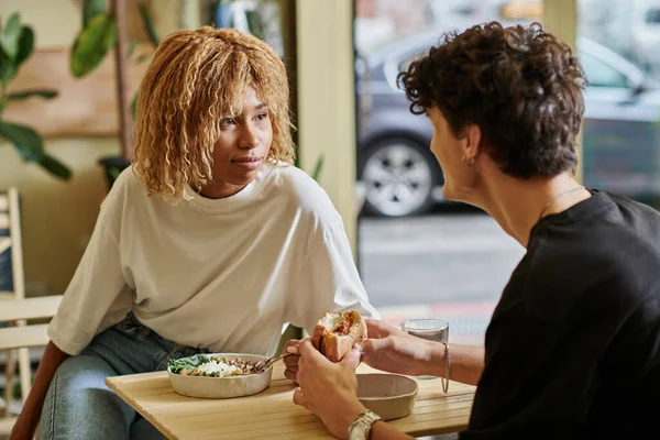 Young african american woman with curly hair eating salad bowl near blurred boyfriend in vegan cafe — Stock Photo