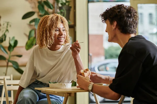 Glücklich afrikanisch-amerikanische Frau mit Zahnspange isst Salatschüssel neben verschwommenem Freund in veganem Café — Stockfoto