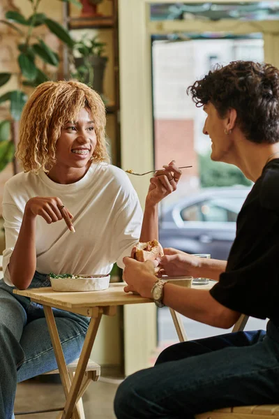 Feliz mulher afro-americana com aparelho comendo salada perto de namorado encaracolado borrado no café vegan — Fotografia de Stock