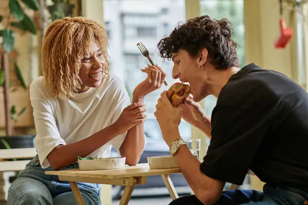 Happy african american woman with braces looking at curly boyfriend eating tofu burger in vegan cafe — Stock Photo