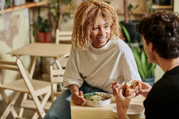 Happy african american woman with braces looking at curly boyfriend eating vegan burger in cafe — Stock Photo