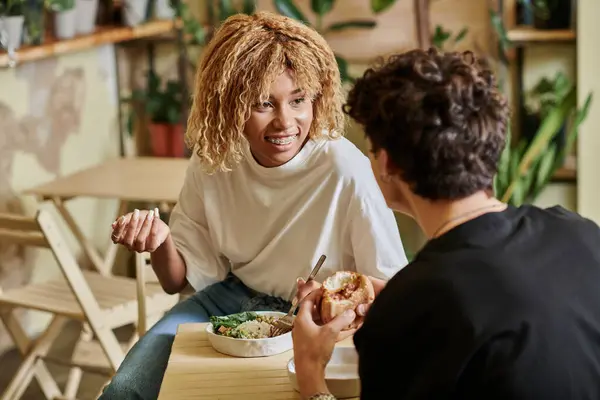 Felice ragazza africana americana con bretelle guardando riccio fidanzato mangiare hamburger vegan in caffè — Foto stock