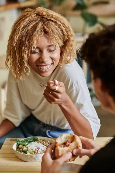 Cheerful african american woman with braces eating salad near curly boyfriend with tofu burger — Stock Photo