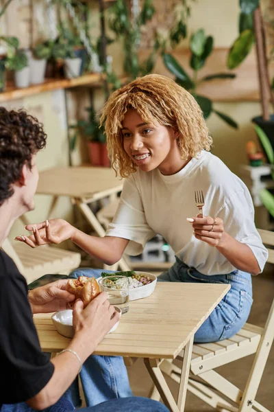 Счастливая африканская американская девушка болтает с кудрявым парнем возле вегетарианской еды в веганском кафе — стоковое фото