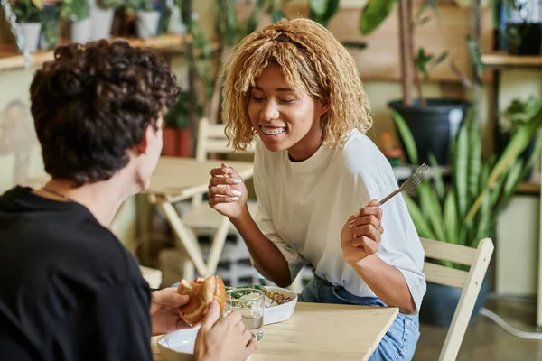 Excited african american girl chatting with curly boyfriend near plant based meal in vegan cafe — Stock Photo