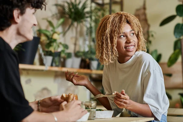 Happy african american girl eating her plant-based meal near curly boyfriend in vegan cafe — Stock Photo