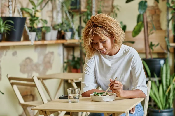 Jovem mulher afro-americana em aparelho comendo salada fresca em tigela dentro de café vegan repleto de plantas — Fotografia de Stock