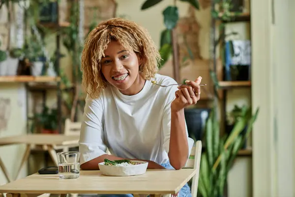 Heureuse femme afro-américaine à bretelles manger de la salade fraîche dans un bol à l'intérieur d'un café végétalien rempli de plantes — Photo de stock