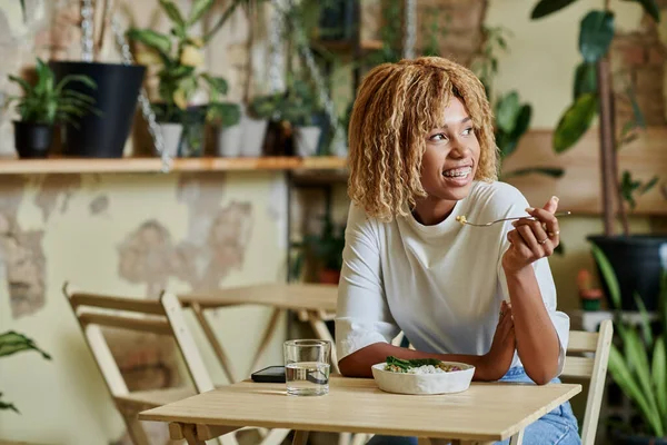 Menina americana africana feliz em aparelhos comendo salada fresca em tigela dentro de café vegan repleto de plantas — Fotografia de Stock