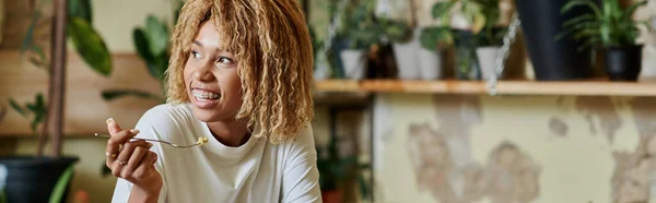 Felice ragazza afro-americana in bretelle mangiare insalata fresca all'interno di caffè vegan pieno di piante, banner — Foto stock