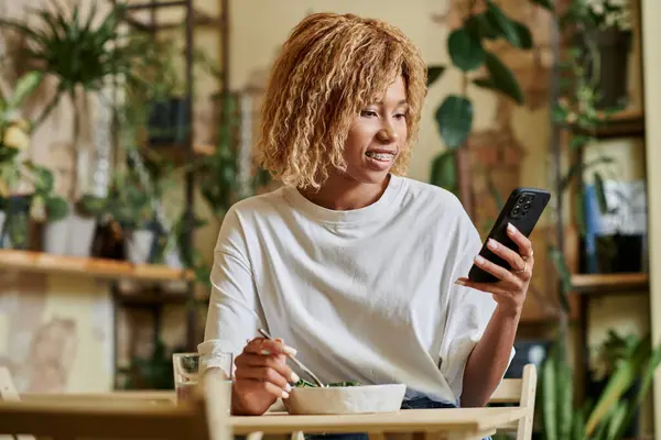 Cheerful african american girl in braces using smartphone and eating fresh vegan salad in cafe — Stock Photo
