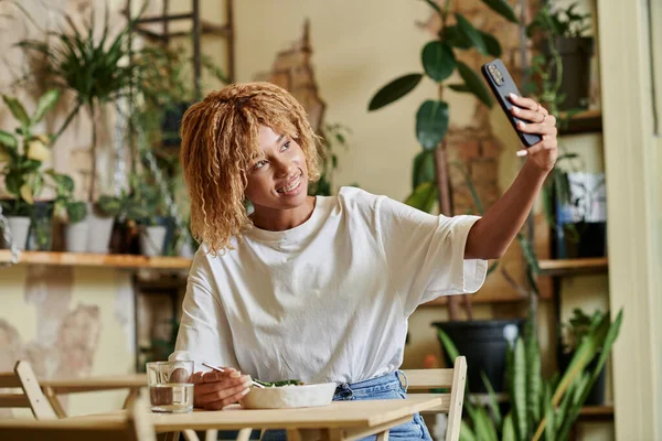 Gai afro-américaine fille dans bretelles prendre selfie tout en mangeant salade végétalienne fraîche dans le café — Photo de stock