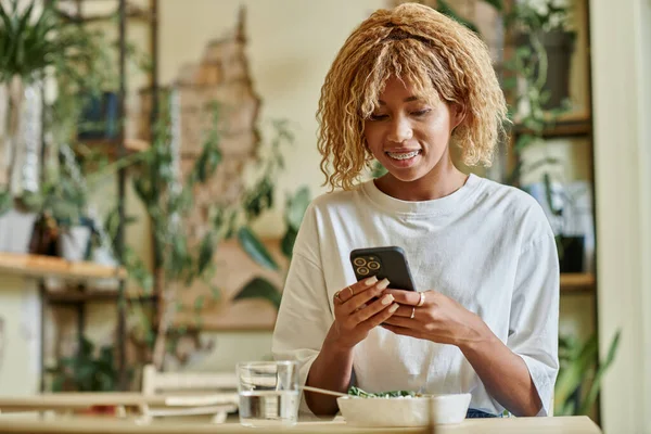 Mulher americana africana feliz em aparelho usando smartphone perto de tigela de salada vegan fresco no café — Fotografia de Stock