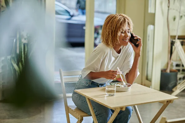 Glückliche junge dunkelhäutige Frau in Hosenträgern, die mit dem Smartphone spricht und im Café frischen veganen Salat isst — Stockfoto