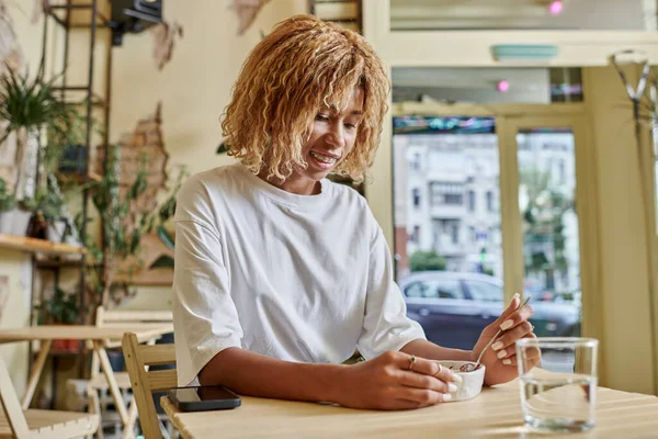 Pleased dark skinned young woman with braces holding fork near fresh vegan salad in modern cafe — Stock Photo