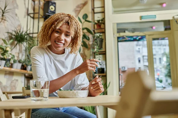 Joyeuse jeune femme à la peau foncée avec bretelles tenant fourchette près de salade végétalienne fraîche dans un café moderne — Photo de stock