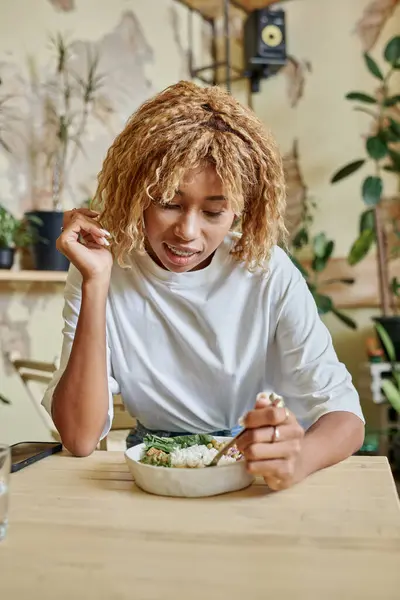 Cheerful dark skinned girl with braces holding fork near fresh vegan salad in modern cafe — Stock Photo