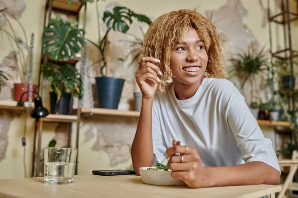 Dreamy dark skinned girl with braces holding fork near fresh salad bowl in modern vegan cafe — Stock Photo