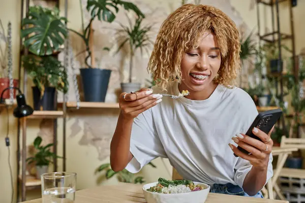 Heureux sombre peau jeune femme avec bretelles tenant fourchette près saladier dans le café végétalien moderne — Photo de stock