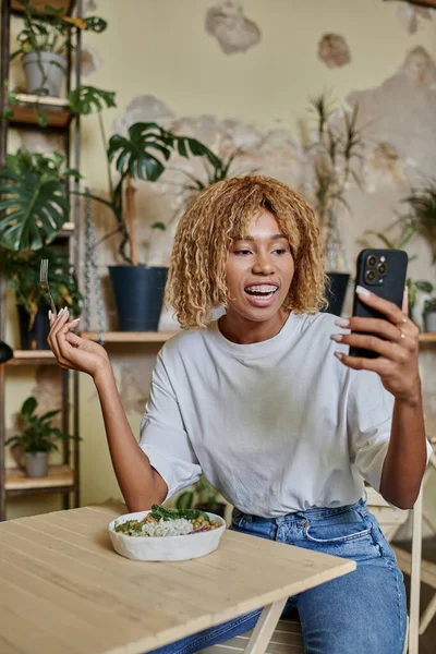 Excité sombre peau jeune femme avec bretelles tenant fourchette près de salade végétalienne et en regardant smartphone — Photo de stock