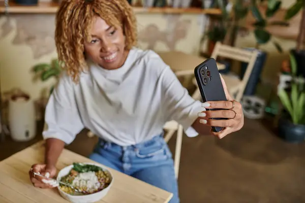 Blurred african american woman taking selfie on smartphone in plant-filled vegan cafe, meal time — Stock Photo