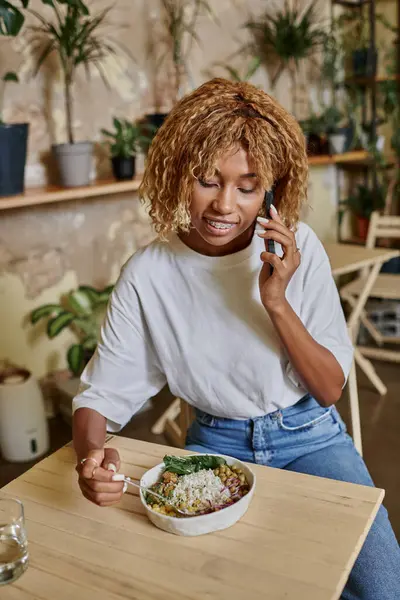 Mujer joven de piel oscura feliz con frenos que sostienen tenedor cerca de ensalada vegana y hablando en el teléfono inteligente - foto de stock