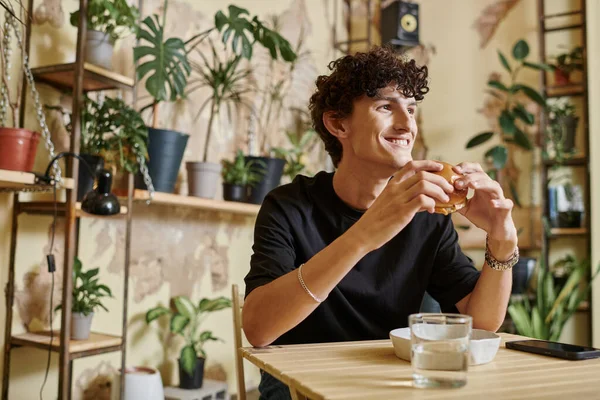 Happy young man holding tofu burger and looking away while smiling in vegan cafe, enjoyment — Stock Photo