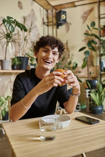 Cheerful young curly man holding plant-based tofu burger and smiling in vegan cafe, enjoyment — Stock Photo