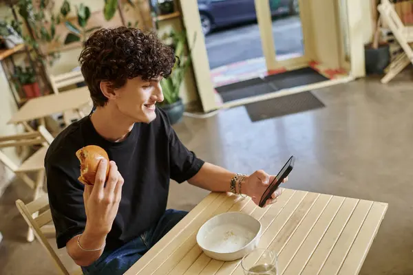 Joyeux jeune homme bouclé tenant hamburger de tofu à base de plantes et en utilisant smartphone dans un café végétalien — Photo de stock