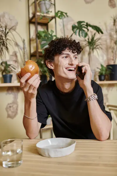 Jeune homme bouclé positif tenant hamburger de tofu à base de plantes et parler sur smartphone dans un café végétalien — Photo de stock