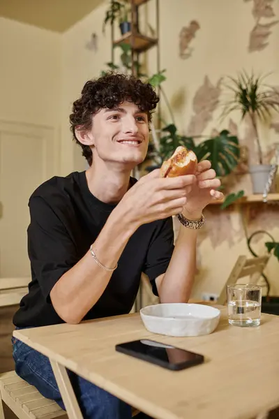 Jeune homme positif et bouclé tenant burger de tofu biologique et souriant dans un café végétalien rempli de plantes — Photo de stock
