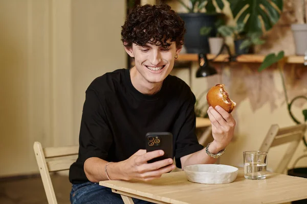 Smiling and curly young man holding plant-based tofu burger texting on smartphone in vegan cafe — Stock Photo