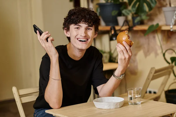 Heureux et frisé jeune homme tenant hamburger de tofu à base de plantes et smartphone dans un café végétalien, la vie de la ville — Photo de stock