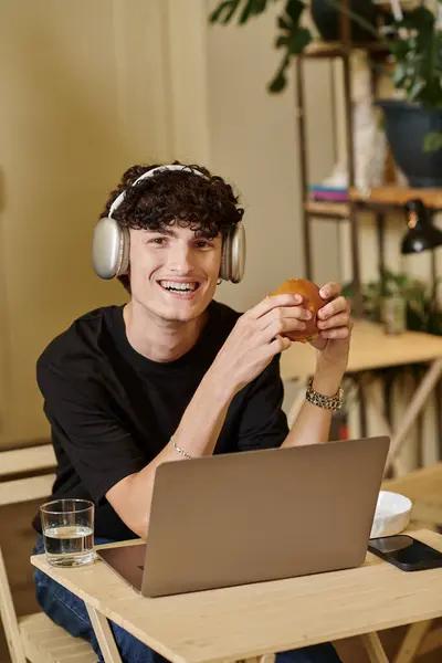 Hombre positivo en auriculares inalámbricos utilizando el ordenador portátil y disfrutar de hamburguesa de tofu en la cafetería vegana, a base de plantas - foto de stock
