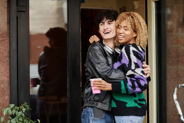Радісна і різноманітна молода пара в повсякденному одязі обіймається біля входу в веганське кафе, дата кави — стокове фото