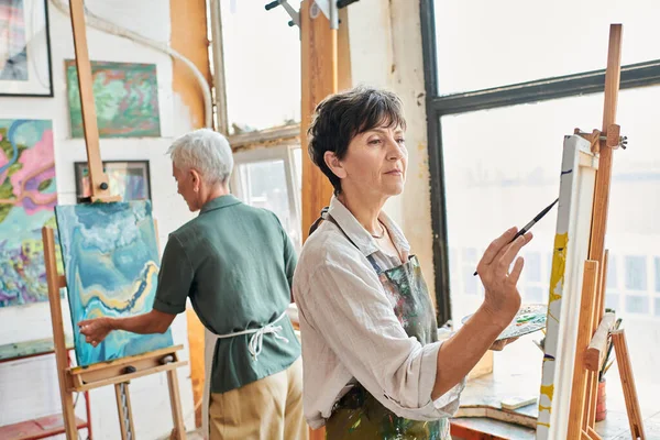 Inspired brunette mature woman painting on easel near female friend in art workshop, creativity — Stock Photo