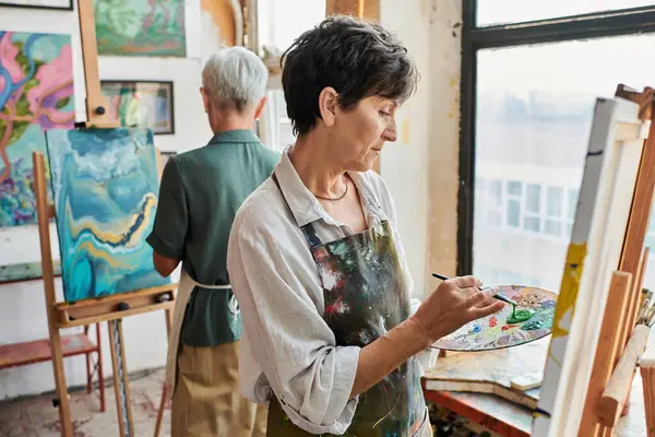 Bruna di talento donna matura pittura su cavalletto vicino amico femminile in laboratorio d'arte, hobby creativo — Foto stock