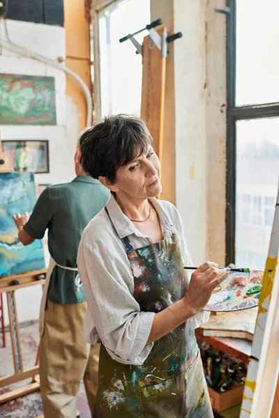 Riflessivo bruna donna matura pittura su cavalletto vicino amico femminile in laboratorio d'arte, creatività — Foto stock