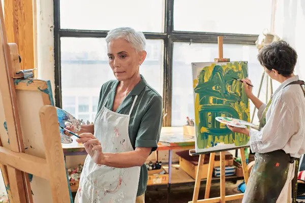 Femme mûre aux cheveux gris dans la peinture de tablier sur chevalet proche amie dans un atelier d'art, créativité — Photo de stock