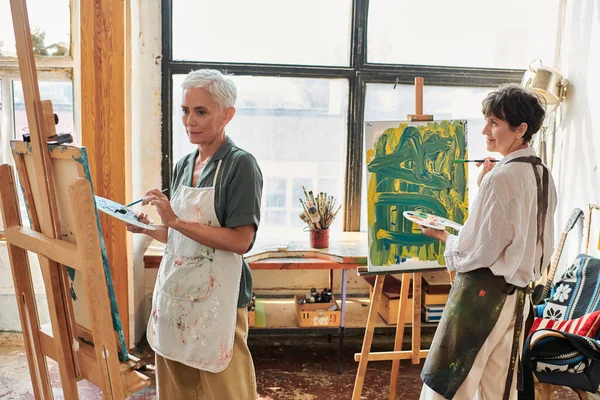 Talented mature female friends in aprons painting on easels in modern art workshop, creative hobby — Stock Photo