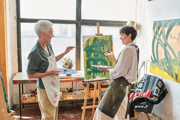 Joyful woman pointing at easel with colorful painting near female friend in art studio, teamwork — Stock Photo