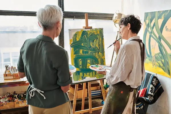Pensive mature woman looking at her painting near female friend in craft workshop, creative hobby — Stock Photo