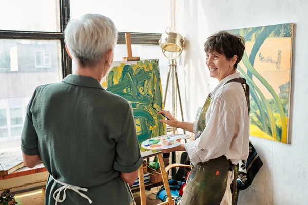 Smiling mature woman pointing at her painting near female friend in craft workshop, creative hobby — Stock Photo