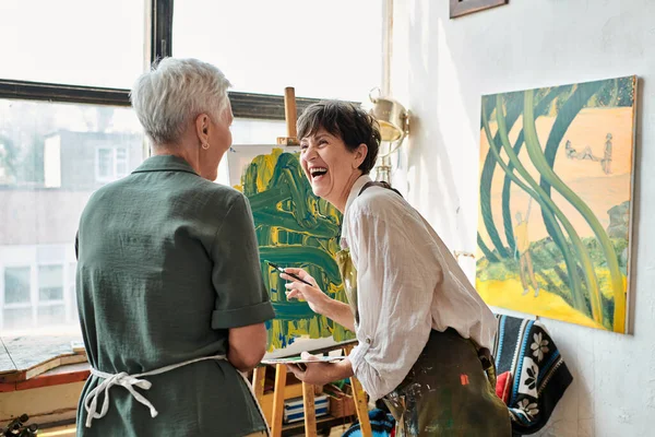 Excited mature woman laughing near easel and  female friend in craft workshop, creative hobby — Stock Photo