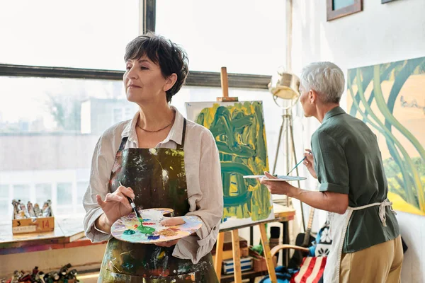Inspired mature woman holding palette while female friend painting in art workshop, creative leisure — Stock Photo