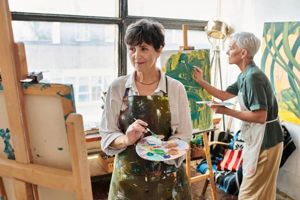 Brunette mature woman looking at easel while female friend painting in art workshop, creative hobby — Stock Photo