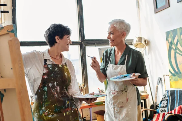 Cheerful mature female artists in aprons holding palettes and talking in modern art workshop — Stock Photo