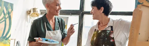 Joyful mature female artists in aprons holding palettes and talking in modern art workshop, banner — Stock Photo